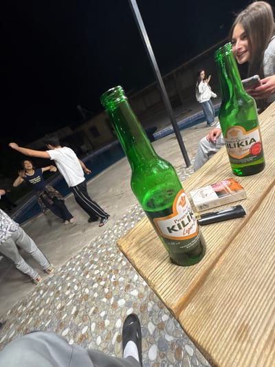 The image shows an outdoor social gathering, likely at night, possibly in Dzoraghbyur, Armenia, but this cannot be verified from the image. The scene includes two people at a wooden table: a woman who is partially visible in the upper left, and another person seated at the bottom right, with one foot visible. On the table are two green glass bottles of "Kilikia" beer, a pack of Winston cigarettes, and a black vape pen. Two additional people are seen lying on the ground in the lower portion of the image, one in a white T-shirt and the other in a blue shirt. There's a dark environment visible in the background, possibly indicating the time of day. The flooring under the table consists of a stone pebble mosaic design.