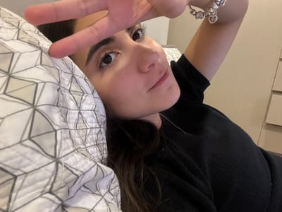 The image displays a young adult female lying in an indoor setting, likely a bedroom. The individual has dark hair and light-to-medium skin, and is looking directly forward. Her head rests on a white pillow featuring a gray geometric pattern of interconnected cubes. She wears a black long-sleeved top and a thin silver-colored necklace. Her right hand is raised, with the index and middle fingers forming a "V" sign, partially obscuring the upper portion of her face. A silver-colored charm bracelet, adorned with multiple charms including a heart, is visible on her right wrist. The background on the right reveals a light-colored wall or furniture piece, possibly a dresser, exhibiting faint horizontal lines. There is no discernible information within the image to specify a location within Santa Catarina, Mexico, or any other geographical area.