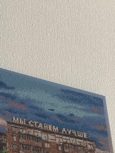 The image shows a textured, off-white wall on the left. On the right side of the frame, there's a diamond painting that features a building with blue sky. There is no indication that this picture was taken in Анкудиновка, Russia.