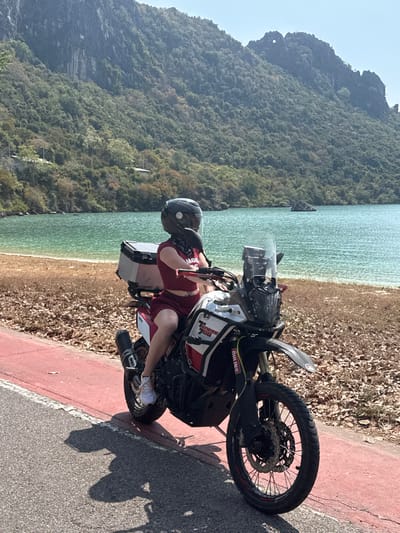 A person wearing a helmet is sitting on a Yamaha Tenere 700 motorcycle on a paved road. Adjacent to the road is a strip of brown vegetation followed by a sandy beach and light blue water. The backdrop features a densely forested mountain range under a clear sky. The motorcycle is facing right and slightly uphill. The scene suggests a coastal or island setting, possibly during a ride stop.