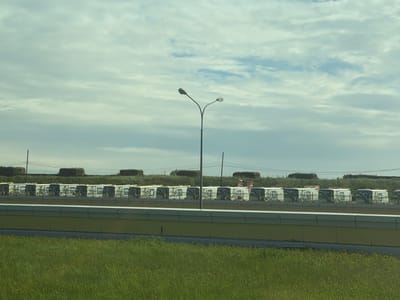 The image depicts a street in Lisy Nos, Russia, with a row of identical small trailers or mobile units parked behind a low concrete barrier painted yellow and gray. Behind the trailers, there are stacks of hay bales and some green foliage. In the foreground, there is a grassy area. A street light stands tall in the center of the frame. The sky is overcast with light and dark clouds. Power lines run across the background.