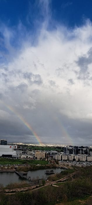 The image, when properly oriented, depicts a panoramic view of a city landscape under a dramatic sky, featuring a prominent rainbow.
The sky is split, with the left side showcasing a clear, vibrant blue with scattered, wispy white clouds. As the eye moves towards the right, the sky becomes increasingly overcast with heavier, grey and white clouds, indicating recent or ongoing precipitation. Arcing across this cloud-filled portion of the sky is a distinct, colorful primary rainbow. A fainter, secondary rainbow is also visible just above the primary one.
Below the sky, a sprawling urban area stretches horizontally across the middle of the frame. Numerous buildings, mostly light-colored and of varying heights, form the cityscape, extending into the distant horizon. To the right of the buildings, a body of water, possibly a river or canal, snakes through the landscape. The water appears somewhat dark and reflective. Lush green vegetation, including trees and bushes, lines the banks of this water body and is also visible within and around the urban structures.
The overall impression is one of a transition in weather, with clear skies giving way to rain, beautifully punctuated by the double rainbow arcing over the urban and natural landscape. The lighting suggests daytime, possibly after a rain shower, with the sun casting light from behind the viewer, illuminating the rainbow. No discernible text is visible in the image.
