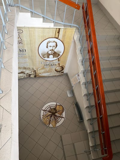 This is an eye-level, indoor shot looking down a stairwell with grey tile steps and brown wooden handrails. The walls adjacent to the stairs are also covered with grey tile arranged in a diamond pattern. A historical mural is visible on the wall, featuring a portrait of Jean Bernard Leon Foucault, his name, and the dates 1819-1868. Additional Russian text related to Foucault is also included in the mural. On the floor, a compass design is visible. The presence of the text "ЛУКОТАЙМ" suggests the location is Murmansk, Russia, where a Foucault pendulum is known to exist.