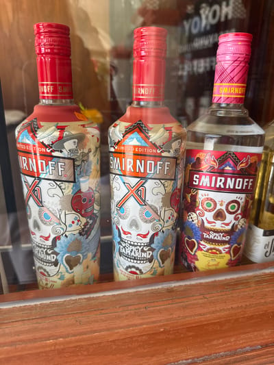 The image shows a close-up view of three bottles of Smirnoff vodka in a display case, possibly a liquor store or bar in San Cristóbal, Mexico. The bottles have colorful Day of the Dead-themed designs on their labels. The top bottle is "Spicy Tamarind" flavor. The other two are a Smirnoff X Limited Edition. The scene is well-lit, and the bottles appear to be new and in good condition. The wooden frame of the display case is visible along the right edge.