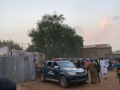 The scene shows a large crowd of people gathered in an open area in Sokoto, Nigeria, at coordinates 13.049, 5.238. There is a large tree to the left and a building to the right. A dark blue pickup truck with the words "Police Strike Force" clearly visible on the side is parked facing the crowd. The truck is facing the camera with its headlights on. There are many people standing behind and to the side of the truck, some looking at the camera and some looking towards the truck. The scene is lit by the sun, which is setting in the background.