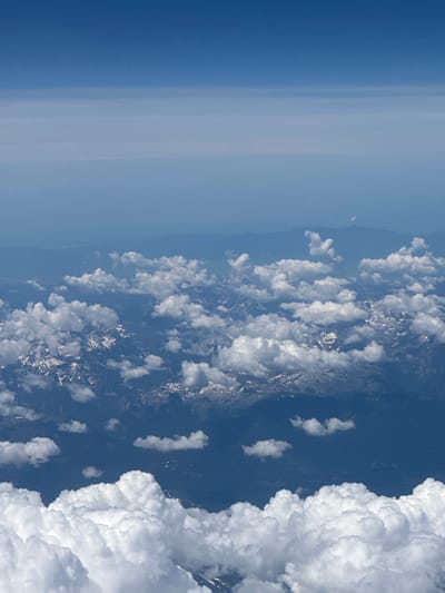 An aerial view captures a vast expanse of the sky and a mountain range below, dotted with clouds. The sky transitions from a deep blue at the top to a lighter, hazy blue towards the horizon. Wisps of clouds are scattered across the upper and middle sections of the image. The mountain range beneath is partially obscured by these clouds, with some peaks showing patches of snow. Dark green foliage covers the lower slopes of the mountains. In the foreground, a large formation of white, fluffy clouds fills the bottom of the frame, casting shadows on the mountains below. The image does not provide specific details about the city of Unknown, Kosovo.