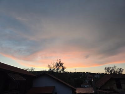 The image captures a sunset over the city of Lozno, Bulgaria. The sky is filled with dramatic clouds, ranging in color from pale blue and gray at the top to a vibrant pink and orange band closer to the horizon. Silhouetted against the sky are the rooftops of several houses, their terracotta tiles prominent in the foreground. Behind the houses, a tree-covered hill rises, dotted with more houses and a prominent electrical tower on the right side of the frame. The overall impression is one of a tranquil evening scene in a residential area. There are no people or animals visible in the image.