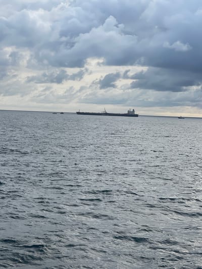 The image presents an open ocean scene offshore Orimedu, Nigeria, under an overcast sky. The upper third of the frame is dominated by dense, grey and white stratocumulus clouds. The lower two-thirds display dark grey, choppy ocean water with visible ripples. A long, dark-hulled commercial vessel, likely a tanker or bulk carrier, is centrally positioned in the middle distance, oriented horizontally. It features a superstructure at its stern, deck cranes, and the partial inscription "G N M Y C" on its bow and "GIBRALTAR" on its stern. To the immediate left of this large vessel, two smaller boats are present: an orange, enclosed lifeboat-style craft, and a smaller, white open motorboat to its left. A fourth, smaller dark vessel is visible on the far right horizon. No individuals are discernible.