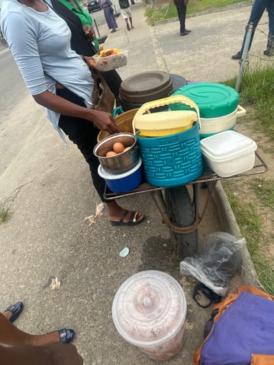 A street vendor stands next to a cart loaded with stacked containers, presumably containing food. The cart's wheel is visible. The vendor is holding a package of food in one hand and a metal bowl with two eggs in the other. Beside the cart is a large bucket with a lid. The area is on the sidewalk. A few other people are visible in the background.