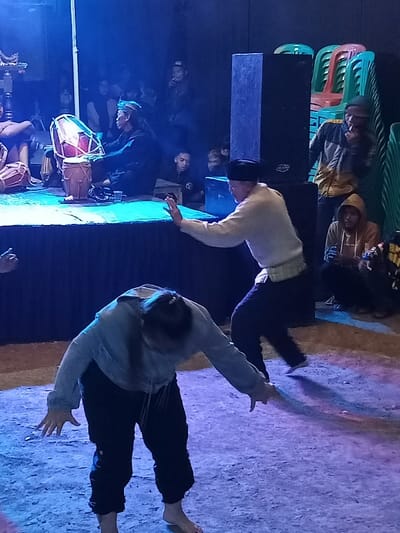 This nighttime image captures a dynamic cultural performance in Pangalengan, Indonesia, under striking blue stage lighting. In the foreground, two individuals are engaged in what appears to be a traditional dance or martial arts demonstration. One performer, wearing a light-colored long-sleeved shirt, dark pants, and a dark cap, is captured in a powerful, open-handed stance with one leg slightly raised. The second performer is bent low, almost parallel to the ground, with arms extended outwards and fingertips nearly touching the ground. This performer is dressed in a grey hooded top and dark pants, and both are barefoot.
On a raised, dark-skirted stage in the background, musicians provide live accompaniment. A central musician, adorned in dark attire and a headwrap, is seated and intensely playing a set of traditional drums, known as kendang. Other figures are partially visible on the stage, suggesting additional performers or musicians.
The scene is set in what appears to be an indoor or sheltered outdoor venue, with artificial blue lights dramatically illuminating the performers and the stage. The ground beneath the performers is a textured, light-colored surface, possibly dirt or concrete, further highlighted by the blue light. Behind the stage, an audience is gathered, observing the performance. Some spectators are standing, while others are seen through the shadows and gaps around large black speaker units and stacks of colorful plastic chairs. One audience member on the right is visible smoking. No legible text is present in the image.