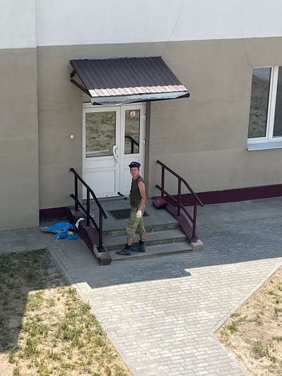 A young man, wearing a camouflage-patterned uniform and a dark blue cap, stands on a paved pathway in front of a building. He is looking directly at the camera with a neutral expression. His attire suggests he might be in some form of military or outdoor service.
The building appears to be an apartment complex or a similar multi-story structure, with a light-colored facade and a dark brown awning over the entrance. The entrance itself is a glass door with a white frame. To the right of the entrance, there are metal railings leading up a few steps, and a blue plastic bag with white contents rests on the ground near the railings.
The pathway in front of the building is paved with light-colored rectangular stones. To the right of the pathway, there is a patch of dry, grassy ground with sparse green vegetation. The lighting indicates it is daytime, with sunlight casting shadows. The overall scene is outdoors, and the location context provided suggests it is in Belarus.