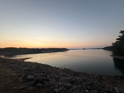 The image captures a serene sunset over Lake Lanier in Buford, Georgia. The sky transitions from a soft orange and yellow glow near the horizon to a pale blue overhead. A calm, expansive body of water stretches towards the distance, reflecting the muted colors of the sky. The shoreline in the foreground is characterized by a rocky embankment with dry grass and scattered vegetation. Dense, dark green trees line the far shore, creating a silhouetted landscape against the fading light. Several small islands or peninsulas are visible in the water. The scene is tranquil and devoid of any human presence or activity.