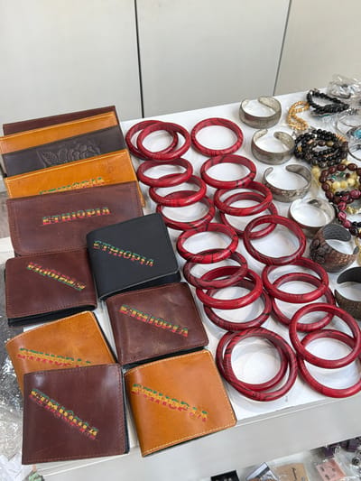 The image depicts a display of goods for sale. A variety of jewelry items, including necklaces, bracelets, and bangles, are positioned on the upper left side of the image. To the lower center, there is a selection of leather wallets in brown and tan colors, each labeled with the word "Ethiopia." The items are arranged on a white surface, likely a table or display case.

This is consistent with a location in Addis Ababa, Ethiopia, which is the origin of the "Ethiopia" labeled wallets, and a common location for tourists to be sold handicrafts.