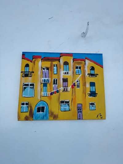 This is a photograph of a painting hanging on a white, textured wall. The painting depicts a yellow building with red roofs and a blue sky. The building has several windows and doors, some with balconies. There is a line of colorful laundry hanging across the middle of the building. The painting is signed in the bottom right corner with the initials "XB". The wall has some minor imperfections, including what appear to be water stains and cracks. There is also a metal hook protruding from the wall above the painting. The photograph is taken at an angle, with the painting slightly off-center. The location in the city of Almaty, Kazakhstan is not discernible from the image itself.
