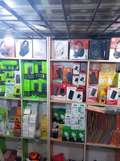 The image shows a shelf in a shop located in Isunba, Nigeria. The shelves are stocked with consumer electronics and accessories including headphones (JBL, AKZ), power banks (Oraimo, Itel), chargers (Itel, Chupez, SHPlus), and phone accessories. The items are arranged in boxes and packages. The shelving unit appears to be made of wood. The lighting in the shop is provided by a fluorescent bulb. In the bottom part of the shelf, there are cables and small storage items.