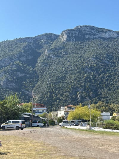The image shows a rocky mountain densely covered with trees, dominating the background. In the foreground, a parking area is visible with a gravelly surface and patches of dry grass. Several vehicles are parked in the area, including a white van with yellow stripes, another white van, a silver car, and a motorcycle. Buildings, possibly residences or businesses, are situated at the base of the mountain, some with red-tiled roofs. Lush green trees and lighter green, leafy trees are interspersed among the buildings and along the edge of the parking area. The sky is clear and blue. The scene appears to be a street or access road within the Municipal Unit of Kamena Vourla, Greece, likely a commercial or residential area at the foot of a mountain.
