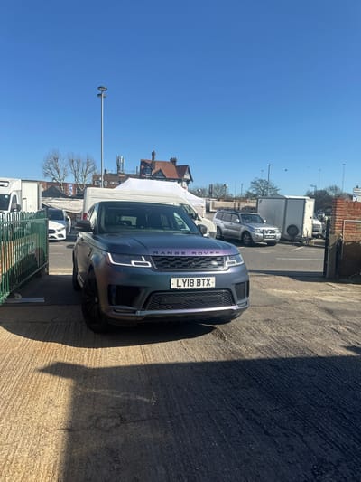The scene depicts an outdoor parking area in Basildon, United Kingdom, specifically identifiable by the distinctive half-timbered building of The Moon on the Square pub located on East Square, visible in the background. A matte iridescent Range Rover Sport, registration LY18 BTX, is the primary subject, centrally positioned on a cracked asphalt ground. The vehicle's chameleon finish displays hues of dark blue, purple, and green, complemented by black multi-spoke alloy wheels and illuminated LED daytime running lights. Strong shadows are cast from the vehicle and an unseen object to the left, indicating bright, direct sunlight.
To the left of the Range Rover, a dark green metal fence is partially visible. In the midground, a market or vendor area is evident, comprising white commercial vans and several white canopy tents. Behind this, a silver SUV equipped with a roof rack and a front bullbar is parked, with a white enclosed trailer positioned to its rear. A white car is partially visible behind the green fence. The background features the aforementioned pub building, a tall grey lamppost, additional urban buildings with roof-mounted antennae, and bare deciduous trees. A partially obscured road sign on the right indicates "Basildon" and "P" for parking. The sky is clear and bright blue. No individuals are visible, and no actions or interactions are discernible within the frame.