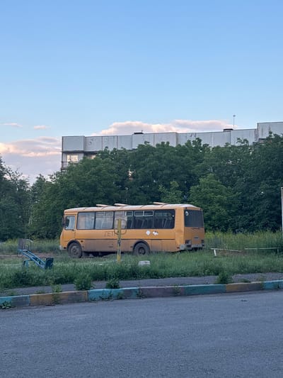 A yellow, dilapidated bus, marked with the Cyrillic letters "ДЕТ", meaning "Children", is parked on a grassy area in Gukovo, Russia. The bus appears abandoned, with one of its windows on the side ajar and various stickers or faded paint markings on its exterior. Behind the bus, a dense line of green trees partially obscures a large, multi-story building with a stark, concrete facade. In the foreground, a section of asphalt is visible, bordered by a low curb painted with alternating colors of yellow, green, and brown. To the left of the bus, a blue and silver seesaw, a common playground item, is situated on the grass. The sky above is a pale blue, with scattered, wispy clouds, suggesting it is either early morning or late afternoon. The overall scene depicts a somewhat neglected urban environment with elements of past community use.