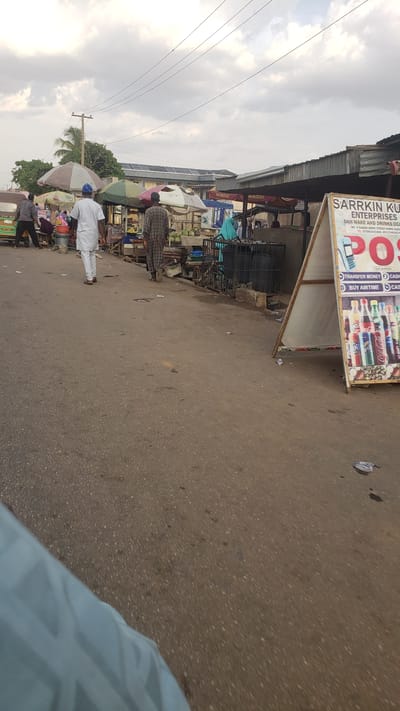 The image shows a street scene in Kawo, Nigeria. A sign for "SARRKIN KU ENTERPRISES" advertises money transfer, airtime, and cash services. Pedestrians walk along the street. Various vendors and shops line the side of the street, sheltered by awnings. The street surface is paved. Overhead electrical wires run along the street. The sky is overcast.
