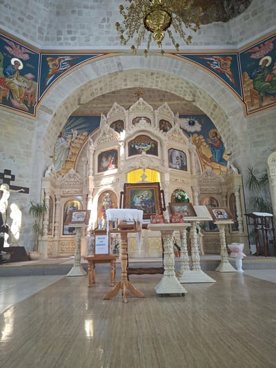 This image depicts the interior of an Orthodox church in Condrița, Moldova. The scene is dominated by an ornate iconostasis, a decorative screen with icons that separates the sanctuary from the nave. The iconostasis is intricately carved and adorned with religious imagery, including depictions of saints and biblical scenes painted in rich colors. Above the iconostasis, a fresco adorns the arched ceiling, featuring more religious artwork and architectural details.
In the foreground, the polished tiled floor reflects the ambient light. Several wooden stands are placed on the floor, holding religious icons and what appears to be a prayer book. A white cloth is draped over one of the stands. A sign with text in Romanian is also visible on one of the stands. To the left of the iconostasis, a large crucifix with a statue of Jesus is mounted on a wall, accompanied by another statue. Potted plants are positioned near the walls, adding a touch of greenery to the scene. A large, ornate chandelier hangs from the ceiling, providing light. The walls are constructed of light-colored stone blocks, consistent with traditional church architecture. The overall impression is one of solemnity and religious devotion.