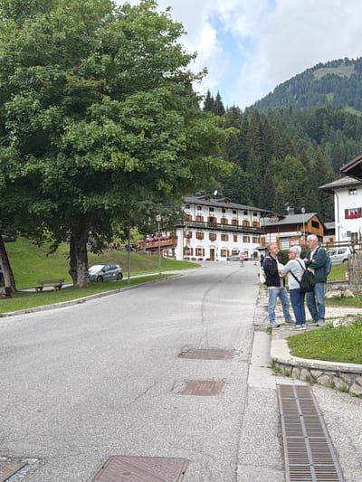 The image depicts a street scene in Sauris di Sopra / Plozn / Sauris Disore, Italy. In the foreground, three individuals, two men and a woman, are engaged in conversation near a street drain and a stone-lined garden bed. The man on the left, wearing a black jacket and blue jeans, faces the other two. The woman, in a light-colored top and blue jeans, stands between the two men, her hands touching the man to her left. The man on the right, wearing a green jacket and blue jeans, looks towards the man on the left.

Further down the street, several buildings line the left side of the road. The most prominent is a multi-story building with white walls, dark shutters, and balconies, suggesting a traditional alpine architectural style. Across the street, a large, leafy tree dominates the left side of the frame, with a wooden bench beneath it. A small, gray car is parked under the tree. In the background, a grassy slope rises behind the buildings, leading to a dense forest of evergreen trees that covers a mountain. The sky above is partly cloudy. The overall impression is a quiet, scenic setting in a mountainous Italian village.