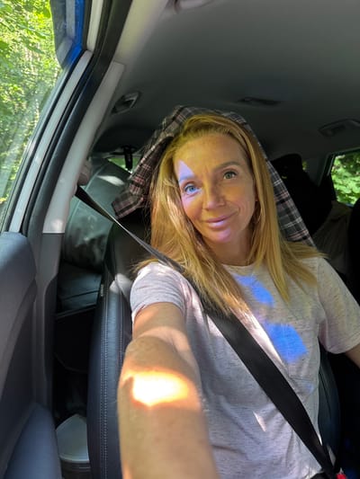 A blonde woman is sitting in the passenger seat of a car, looking at the camera. Sunlight streams through the windshield, casting colorful patterns on her face and shirt. She is wearing a pink t-shirt and has a plaid blanket draped over her head. The car appears to be parked on a road surrounded by trees. The location is the village of Portovoe, Russia.