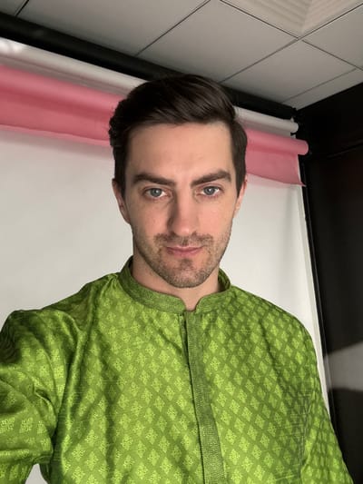 A man with light skin and brown hair is wearing a green, patterned shirt. He has blue eyes and a short beard. He is positioned in front of a white wall with a pink backdrop on the left. There is a black structure visible above the wall. The photograph's location in Ghaziabad, India, cannot be confirmed.