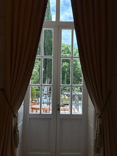 The image shows a view through a pair of French doors, framed by heavy, tan curtains. The doors are divided into multiple panes, revealing an outdoor scene. Through the upper panes, a bright blue sky with scattered clouds is visible, bordered by the tall, dark green foliage of cypress trees. The lower panes offer a glimpse of a garden area. A wooden chair and a small round table are situated on a patio or balcony. Beyond this, a railing with decorative metalwork is visible, separating the seating area from a grassy lawn or park. The overall impression is of an elegant interior looking out onto a manicured outdoor space. The scene appears to be located in Mukhrani, Georgia. There are no people or animals visible in the image.