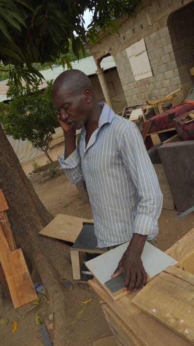An outdoor scene in Masaka, Nigeria, depicts a man engaged in a manual task during bright daylight. The man, with dark skin and a short buzz cut, wears a light blue and yellow striped long-sleeved shirt. He is leaning over a wooden structure, carefully pressing a small, dark gray rectangular object against a white panel with his right hand. His expression appears focused or pensive, with his eyes partially closed as he concentrates on his work.
The setting is characterized by a dirt ground and a mix of construction materials and household items. Wooden planks and beams are prominent, forming a makeshift workbench or structure. In the background, there are concrete block walls of a building, possibly under construction or renovation. Red text, partially legible as "SLEE..." and "MEAS" or "MARS," is visible on one of the concrete blocks, suggesting construction markings. Scattered around are a plastic bottle, yellow leaves, and various pieces of furniture, including a brown sofa and another item with red patterned fabric. Green foliage from trees or bushes frames the left side of the image, adding a natural element to the otherwise functional space. The bright, even lighting indicates clear weather during the day.
