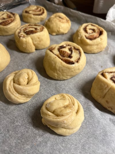 This image captures a close-up, elevated view of ten individual pieces of unbaked dough arranged on a sheet of light gray parchment paper. Six of the dough pieces are light beige, cylindrical rolls with a visible dark brown, granular filling spiraling from their centers, indicative of cinnamon rolls. Two other pieces of dough are intricately twisted or knotted in shape, also light beige. Additionally, one simple, rounded dough portion is partially visible on the far left, and another indistinct, round piece is present in the blurred background. The parchment paper rests on a darker, unidentifiable surface. The background itself is out of focus, showing only indistinct lighter and darker areas. No people are visible in the scene. The location is Schmittweiler, Germany.