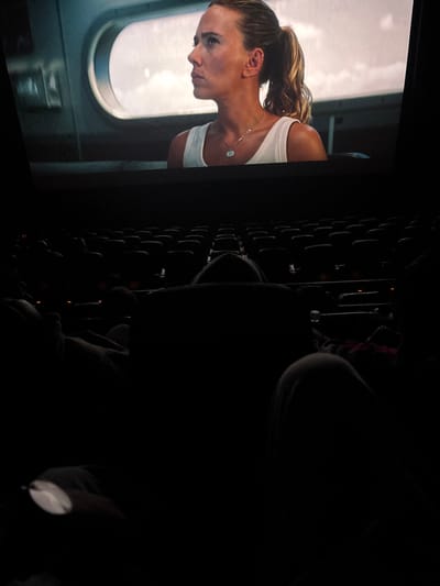A woman in a white tank top looks out of a spacecraft window with a ponytail. A silver necklace adorns her neck. The view outside the window is bright and white. The image seems to be taken inside a dark movie theatre with rows of empty seats visible on the right. Most of the seats are obscured by shadow, highlighting the cinematic setting of the scene.