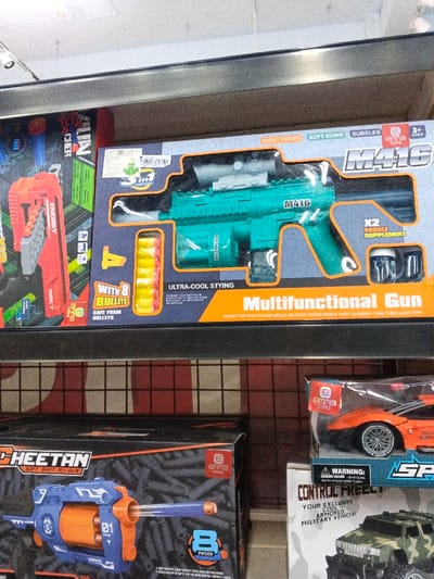 The image captures a section of a retail shelf, likely in a toy store or a department store in Sindanglaya, Indonesia, displaying various children's toys. No people are visible.
On the top shelf, two toy gun packages are prominently displayed. To the left, a red and black package shows part of a red and orange toy gun, with the word "ENERGY" visible. To its right is a larger blue and orange package featuring a teal-green toy M416 rifle with a scope. The packaging highlights "M416," "Multifunctional Gun," and specifies its features: "EIGHT TONES," "SOFT BOMB," "BUBBLES," "ULTRA-COOL STYLING," and "3+ AGES." It includes "WITH 8 BULLETS" of the "SAFE FOAM BULLETS" type and "X2 BUBBLE SUPPLEMENT." The brand "emma TOYS" is visible, along with an Indonesian SNI certification label. Text on the box encourages, "ENJOY THE EAR-TO-EAR SMILE ON KIDS' FACES WHEN THEY UNWRAP THIS THRILLING TOY!"
The bottom shelf presents more toys. On the left, a dark-colored box for a toy gun named "CHEETAN SOFT SHOT TOY GUN" displays a blue and orange revolver-style toy. "emma TOYS" and "8 PIECES" are also visible on this package. To the right, an orange toy car in clear packaging, also from "emma TOYS," sits above a box containing a green toy military vehicle. Text on the military vehicle's packaging reads "CONTROL FREELY," "YOUR EXCLUSIVE COOL ARMORED MILITARY VEHICLE," and includes a "WARNING: CHOKING HAZARD" for children under 3 years. The overall scene suggests a vibrant array of recreational items for children.