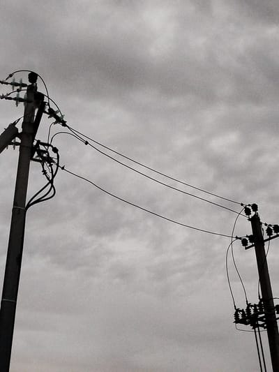 The image shows two utility poles against a cloudy sky. The pole on the left is closer to the foreground and is taller than the pole on the right. Both poles have ceramic insulators and power lines strung between them. The sky is overcast and appears to be a dull gray. The overall mood of the image is somber and somewhat desolate, common for the overcast weather often experienced in the Крупіцкі сельскі Савет region of Belarus. There are no people or animals visible in the frame. The scene appears to be an outdoor, rural or semi-rural setting given the presence of utility poles and the lack of dense urban infrastructure.