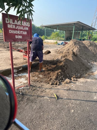 The image depicts a man in a blue head covering and shorts working with soil in front of a red sign with white text in Indonesian. The sign reads "DILARANG BERJUALAN DI SEKITAR SINI," which translates to "NO SELLING AROUND HERE." The man is using a shovel to transfer soil into a red wheelbarrow. Behind him, there is a large pile of soil, and further in the background, a covered structure with green metal framing and a metal roof is visible, possibly a workshop or storage area. The scene appears to be outdoors in a somewhat unkempt area, with dirt, gravel, and some vegetation visible. The overall impression is of a construction or landscaping site. The location is identified as Sugih Waras, Indonesia.