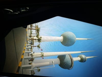 The image shows a side view of the Kuwait Towers, which are a group of three stainless steel towers in Kuwait City, Kuwait. The towers are located on an inlet of the Persian Gulf. The image is taken from a slightly elevated position, possibly from a vehicle, looking towards the towers. The foreground features a road with yellow and black striped barriers and palm trees lining the side. The towers are tall and slender, with spherical upper sections covered in a mosaic of blue and green tiles. They are topped with pointed spires. The sky is a clear blue. The reflection of the towers is visible on a surface, possibly water or a screen, creating a mirrored effect. The image appears to be a photograph or screenshot, possibly of a digital display, as indicated by the presence of what seems like a screen edge at the top and bottom of the frame and a slight pixelation or screen effect. The location of Golbembal, Nigeria is not represented in this image.