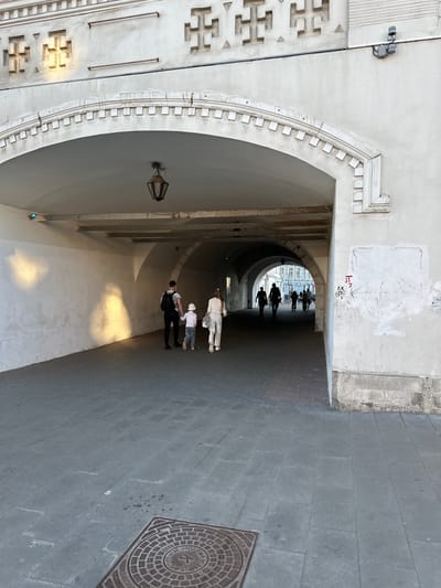 The image shows a view looking through an archway into a tunnel. The archway is made of white stone with decorative elements. Inside the tunnel, there are several people walking. In the foreground, a man, a child, and a woman are walking away from the camera. The man is wearing a black shirt with a pattern and a backpack. The child is wearing a white hat and gray pants. The woman is wearing white clothing. The tunnel walls are white and illuminated by sunlight casting shadows. Further into the tunnel, several silhouettes of people are visible, walking towards the viewer. The ground in the tunnel is paved with dark stones. In the foreground of the image, outside the tunnel, is a manhole cover with circular patterns. The overall environment suggests an urban setting.