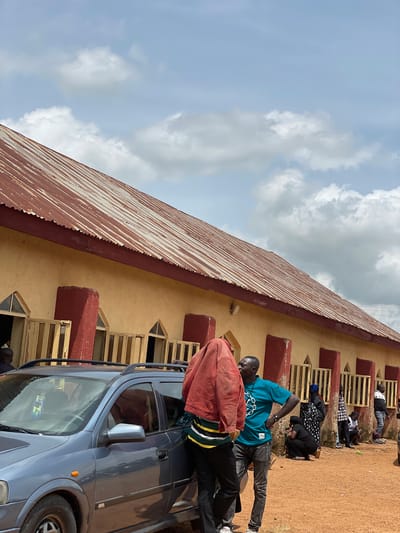 The image depicts an outdoor scene in Ban, Nigeria, in front of a long, tan-colored building with a corrugated metal roof that appears weathered and rusty. The building has numerous windows with yellow wooden slatted coverings and triangular window tops, punctuated by wide, red-painted structural supports. A light blue hatchback car with a roof rack is parked in the foreground to the left.

Several individuals are present. In the foreground on the left, one person is partially obscured by a red jacket draped over their head, wearing a striped shirt and dark pants. To their right, a man in a teal t-shirt and gray jeans stands with his hands on his hips, facing away from the camera. Behind them, several other people are visible. A person in a black and white polka dot garment is crouched near the building. Further down, a person in a plaid shirt stands, and another person is seated on the ground. Near the right edge of the frame, two men stand, one in a black and white patterned shirt and dark pants, and another further in the background wearing a dark shirt and blue jeans. The ground is covered in reddish-brown dirt and sparse dry grass. The sky above is partly cloudy with white and gray cumulus clouds against a pale blue sky.