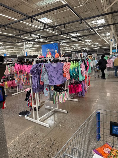 The image displays an interior view of a retail store aisle, primarily featuring children's clothing. The perspective is from eye-level, looking down a long aisle lined with white metal clothing racks.

In the foreground to the right, a metal shopping cart is partially visible, containing bags of what appear to be carrots or other packaged produce. A blue sign attached to the cart clearly reads "Attention:".

The aisle is filled with racks showcasing a wide array of colorful children's apparel, including tops, shorts, and possibly dresses. Many items feature bright hues like pink, purple, teal, and lime green, with various patterns. Notable patterns include purple tops with white butterflies, and a red/pink top with strawberry motifs, sporting a "Wonder" brand tag. Some items are neatly folded on shelves, while others hang from hangers. A few articles of clothing are visible on the speckled, polished concrete floor beneath the racks.

Further down the aisle, a person dressed in a dark jacket and pants is walking away from the camera. Other shoppers are faintly visible in the background. The store boasts a high ceiling with exposed white metal trusses and track lighting, along with several skylights, indicating a daytime setting.

Signs above the clothing displays provide categorization, with visible text including "Casual," "Men's Apparel," and a partially obscured "New! Shoes." Large visual displays featuring mannequins wearing clothing are mounted on the walls behind the racks. The overall scene suggests a busy shopping environment within a large retail establishment.