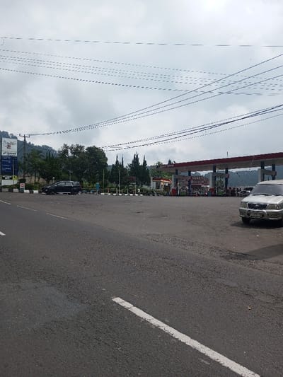 A gas station is visible in the image, with multiple fuel pumps under a red roof structure. A silver SUV is parked to the right of the pumps, while a black SUV is parked further to the left. A signpost is on the far left side of the image. The foreground shows a stretch of asphalt road with a white dividing line. Above the station, a cloudy sky and power lines are evident. Trees and some hills are visible as a backdrop behind the gas station.