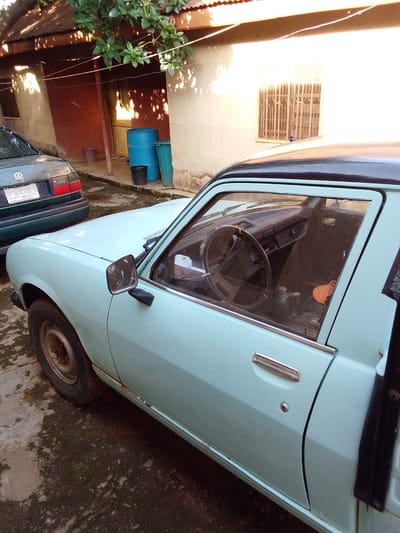 This is a high-angle medium shot of a light blue sedan parked on a concrete surface in Mangu, Nigeria. The car is positioned in front of a building with an orange-colored wall and a corrugated metal roof. The building has a barred window and a door. To the left of the sedan, a dark green Volkswagen sedan is parked. In the background, a blue barrel, a green barrel, and a black bucket are visible near the building. The ground appears to be dirt and concrete. Sunlight casts shadows from the foliage above onto the building and the cars. The blue sedan's driver's side door is ajar, revealing the interior with a steering wheel, dashboard, and passenger seat. The car exhibits some rust along the bottom edge of the door.