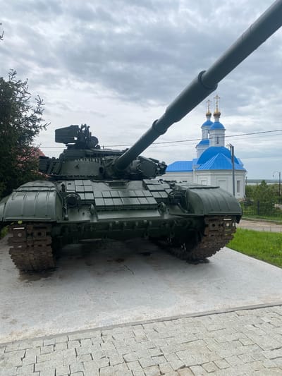 A green military tank, featuring a long cannon, sits on a concrete platform. Behind it, a white church with blue roofs and gold crosses is visible. The sky is overcast with gray clouds. The tank is surrounded by foliage. The ground near the platform is covered in cobblestones. The location is Введенская Слобода, Russia.