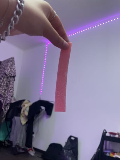 A person's hand, adorned with a silver chain bracelet and two rings, is holding a pink, sugary candy stick. The background of the image is a room illuminated by purple LED strip lights, which are strung along the top of the wall. The room appears to be a bedroom or dorm room, with clothing items, including a leopard print garment, a yellow shirt, and black pants, draped over furniture. In the lower right corner of the image, a black trash bag and a black shelf unit are visible. The overall scene suggests a casual, indoor setting, potentially a private living space. No specific location in Borisova Griva Massiv, Russia can be determined from the visual information.