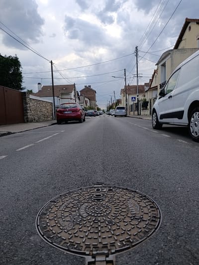 The image shows a low-angle view of a residential street in Villeneuve-Saint-Georges, France, under a cloudy sky. In the foreground, a circular manhole cover is prominent on the asphalt road. The street is lined with houses on both sides, with a stone wall and a wooden gate on the left. Several parked cars are visible on the street, including a red Peugeot on the left and a white van on the right. Power lines and poles are strung across the sky. The overall impression is of a quiet, suburban neighborhood.