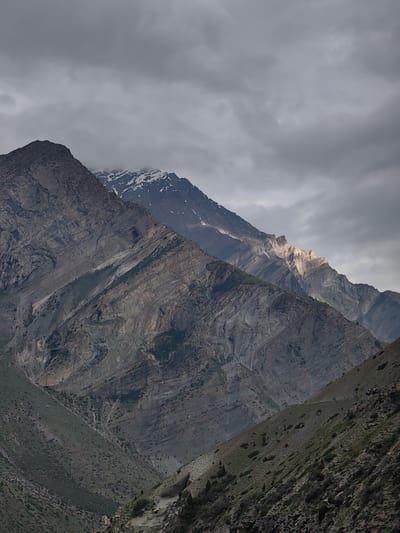 The image shows a dramatic mountain landscape with cloudy skies. The foreground features a steep, rocky slope covered in sparse vegetation. Behind this, a massive mountain range dominates the scene, with intricate rock formations and sharp ridges. Snow can be seen on the peaks of the farthest mountains, suggesting a high altitude. The lighting is dim and moody due to the heavy cloud cover, with shafts of sunlight breaking through in places, illuminating the distant peaks. The image appears to be a landscape shot taken in a mountainous region, likely in the Himalayas, possibly near Gondhla, India, given the visual characteristics of the terrain and vegetation. There are no people or animals visible in the image.