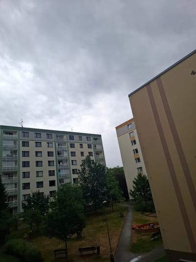 The image shows a residential area with two apartment buildings in Ústí nad Orlicí, Czechia. The sky is overcast. In the foreground, a tall, light brown building with vertical darker brown stripes is angled to the right. To the left, a long, light-colored apartment building with green accents around the balconies and windows is visible. This building has numerous windows and balconies. Between the buildings, trees and a grassy area are present. Further in the distance, another apartment building with a light gray and orange facade is visible. The ground level appears to include a pathway, a sandbox with toys, benches, and a trash can. The overall scene depicts an outdoor view of a multi-story housing complex.