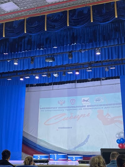 The image depicts an indoor event hall in Tayshet, Russia, specifically focusing on a stage and a portion of the audience. The upper half of the frame features dark blue velvet stage curtains, arranged with draped valances and vertical gold trim at the top, followed by pleated curtains below. A black metal truss system, equipped with multiple stage lights (some emitting purple light) and a projector, hangs in front of the curtains.
The lower half of the frame is dominated by a large white projection screen displaying event information in Cyrillic. The text reads "МЕЖРЕГИОНАЛЬНЫЕ СОРЕВНОВАНИЯ" (Interregional Competitions) "ПО СПОРТИВНОМУ ТУРИЗМУ НА ПЕШЕХОДНЫХ ДИСТАНЦИЯХ" (In Sports Tourism on Hiking Distances), with the prominent stylized word "Сибирь" (Siberia) and "ТАЙШЕТ" (Tayshet) at the bottom. Several logos, including the Russian coat of arms, are visible at the top of the screen. Graphics include a silhouette of a person rappelling on the right and an abstract dark silhouette, possibly a landform, at the bottom left.
Below the screen, the stage is decorated with draped fabric in red, white, and blue. Three white rectangular podiums, trimmed in blue and red, are arranged on the stage floor, numbered "2" (left), "1" (center), and "3" (right). A cluster of red, white, and blue balloons is positioned on the right side of the stage. A dark speaker or equipment box is also visible on the right. In the foreground, the backs of several audience members are visible, seated in dark chairs and observing the stage.