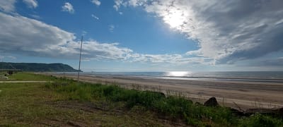 The image depicts a wide, sandy beach with gentle waves lapping at the shore under a bright, partly cloudy sky. The sun is visible, creating a shimmering reflection on the water. In the background, a lush, green hill slopes down towards the coast, and a small cluster of buildings is visible on its lower slopes. The foreground features a grassy area with some scattered rocks and a tall, slender pole. The overall scene suggests a peaceful and natural environment, likely a coastal area in the city of Aleksandrovsk-Sakhalinskiy, Russia, known for its scenic beaches and hilly terrain.