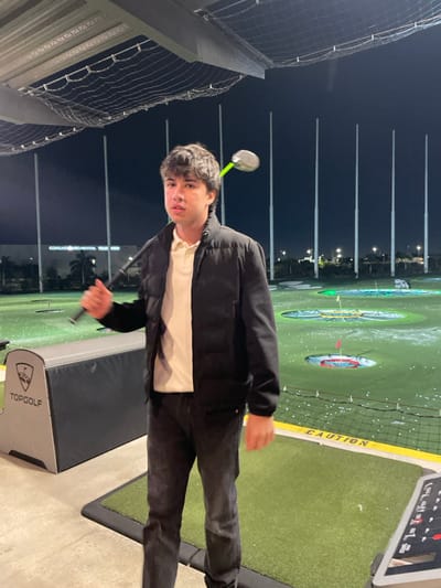 At Topgolf, a young man, dressed in a black jacket over a white shirt and black pants, is about to swing a golf club. The background shows the Topgolf course at night, with targets visible in the distance. Tall net poles line the left side of the frame, and a Topgolf bay divider is in the foreground. The image has been rotated, placing the subject at an angle.
