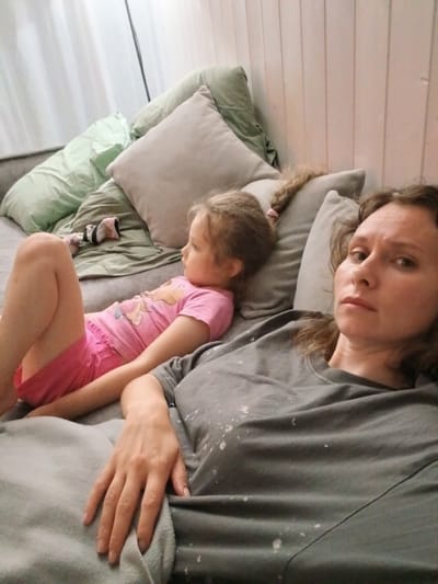 In a dimly lit room, possibly in Verkhneye Dubrovo, Russia, a young girl with blonde hair in a braid, wearing a pink t-shirt and shorts, lies on a grey sofa. She is looking to her left with a thoughtful expression. To her right, an adult woman with brown hair and a grey t-shirt is lying on the sofa, looking directly at the camera with a somber expression. Her hand is resting on her lap. The sofa is covered with several pillows and a grey blanket. The background features vertical wooden panels and white curtains. There are also two pairs of socks visible on the sofa. The overall scene suggests a moment of rest or quiet observation within a domestic setting.