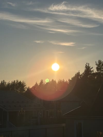 The image shows a sunset view in Ветеран Снт, Russia. The sun is the brightest object in the sky, with a lens flare effect creating a circular halo. Streaky cirrus clouds are visible in the left part of the sky. The silhouette of trees and a two-story house is visible on the right side of the image, obscuring part of the sun. A balcony with railings is visible on the house.